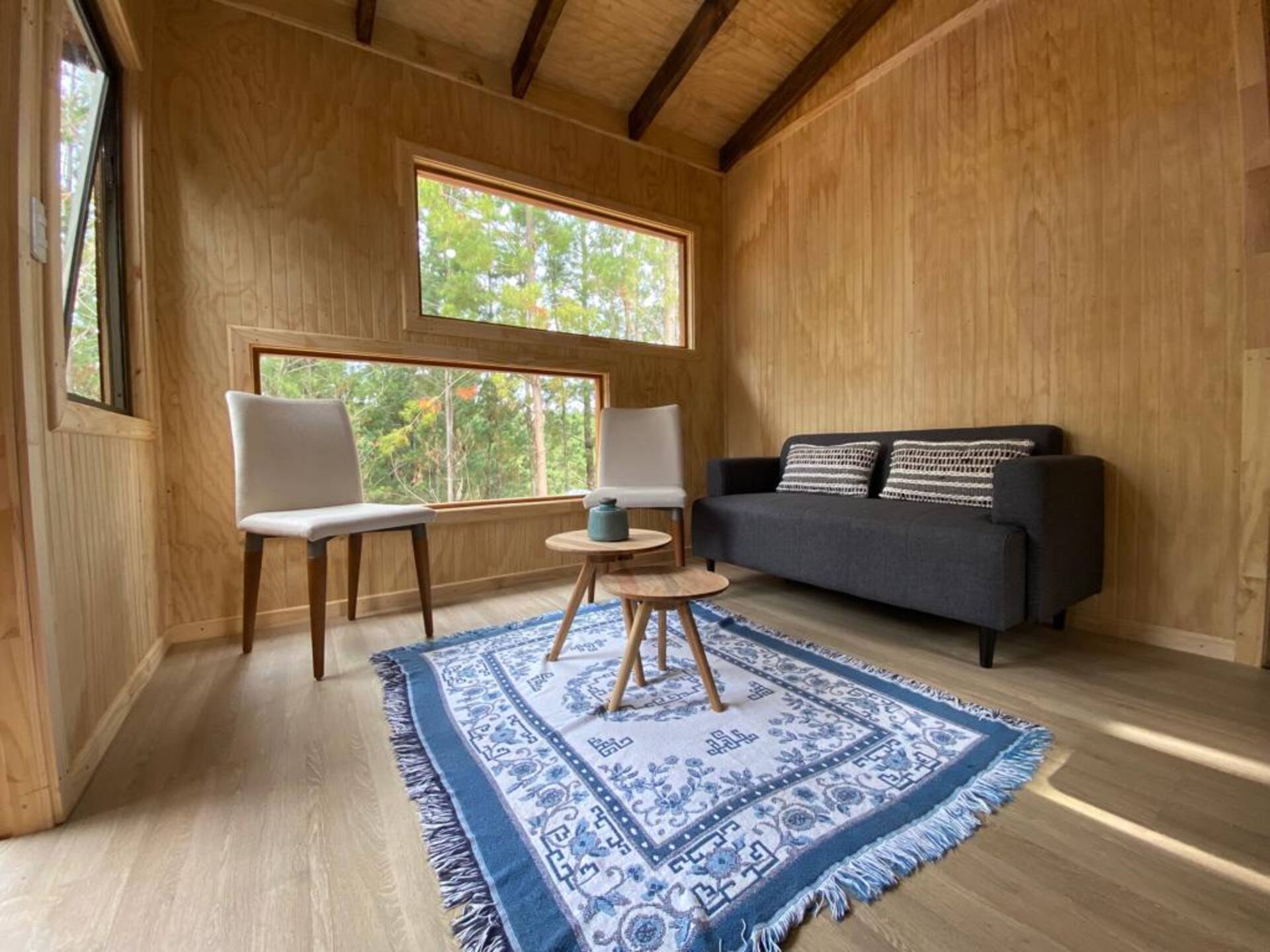 Living de la cabaña con sofá, dos sillas, mesa baja y un ventanal que mira al bosque